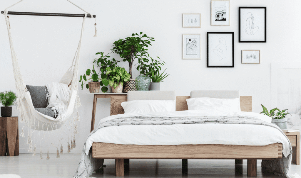 Bright minimalist bedroom with a light wood bed, white bedding, a hanging chair, and a cluster of leafy potted plants on a bench and nightstand creating a calm relaxing atmosphere