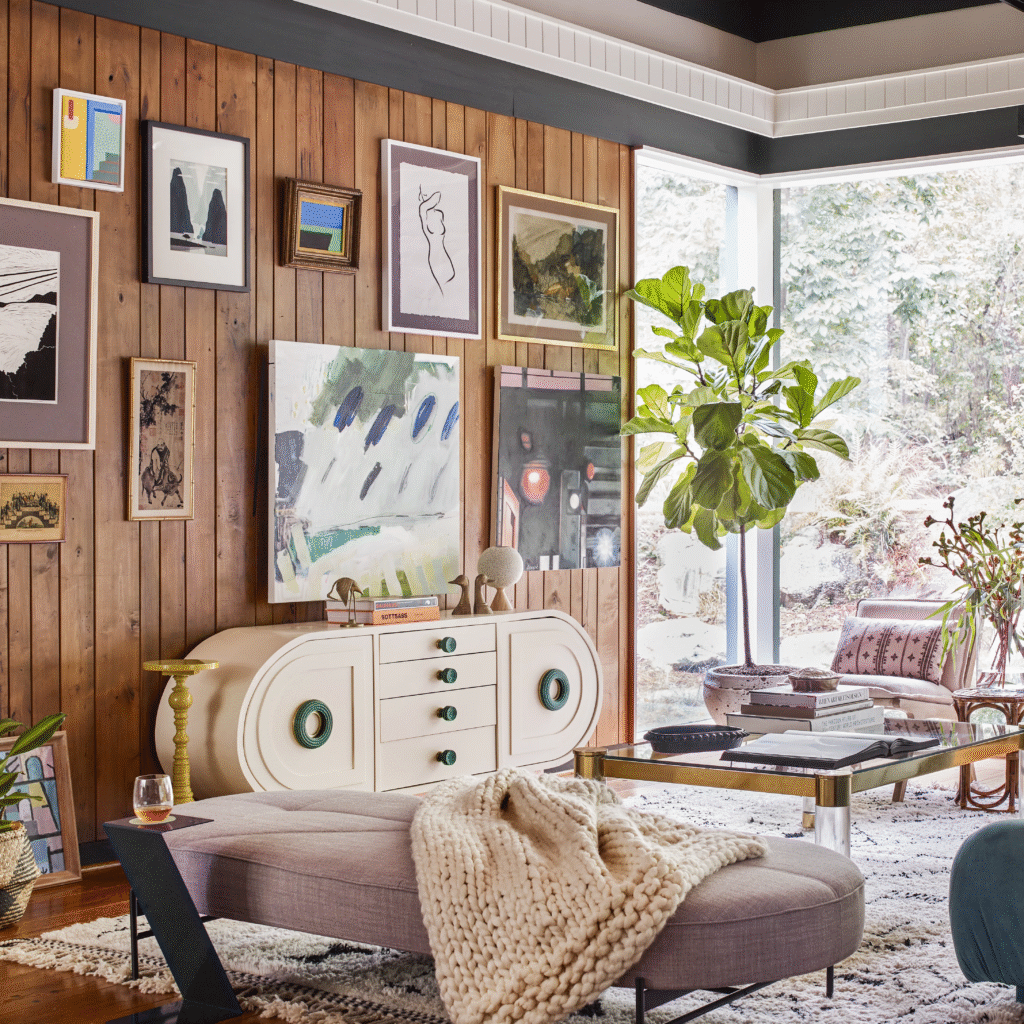 Cozy eclectic living room with wood paneled wall, layered DIY style artwork in mismatched frames above a vintage cream sideboard, large fiddle leaf fig by a picture window, and a bench with a chunky knit throw.