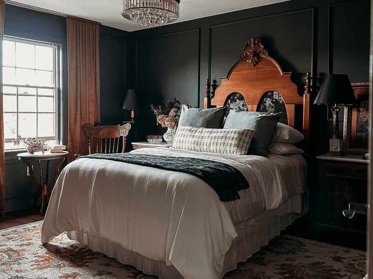 Moody bedroom with dark walls and a carved antique wooden headboard, layered white bedding, vintage side tables, and warm drapes.