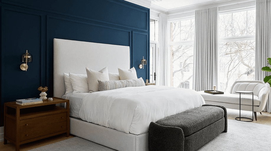 Modern bedroom with a deep navy feature wall, tall upholstered headboard, crisp white bedding, wooden nightstand, and charcoal bench in front of large bright windows with light curtains.