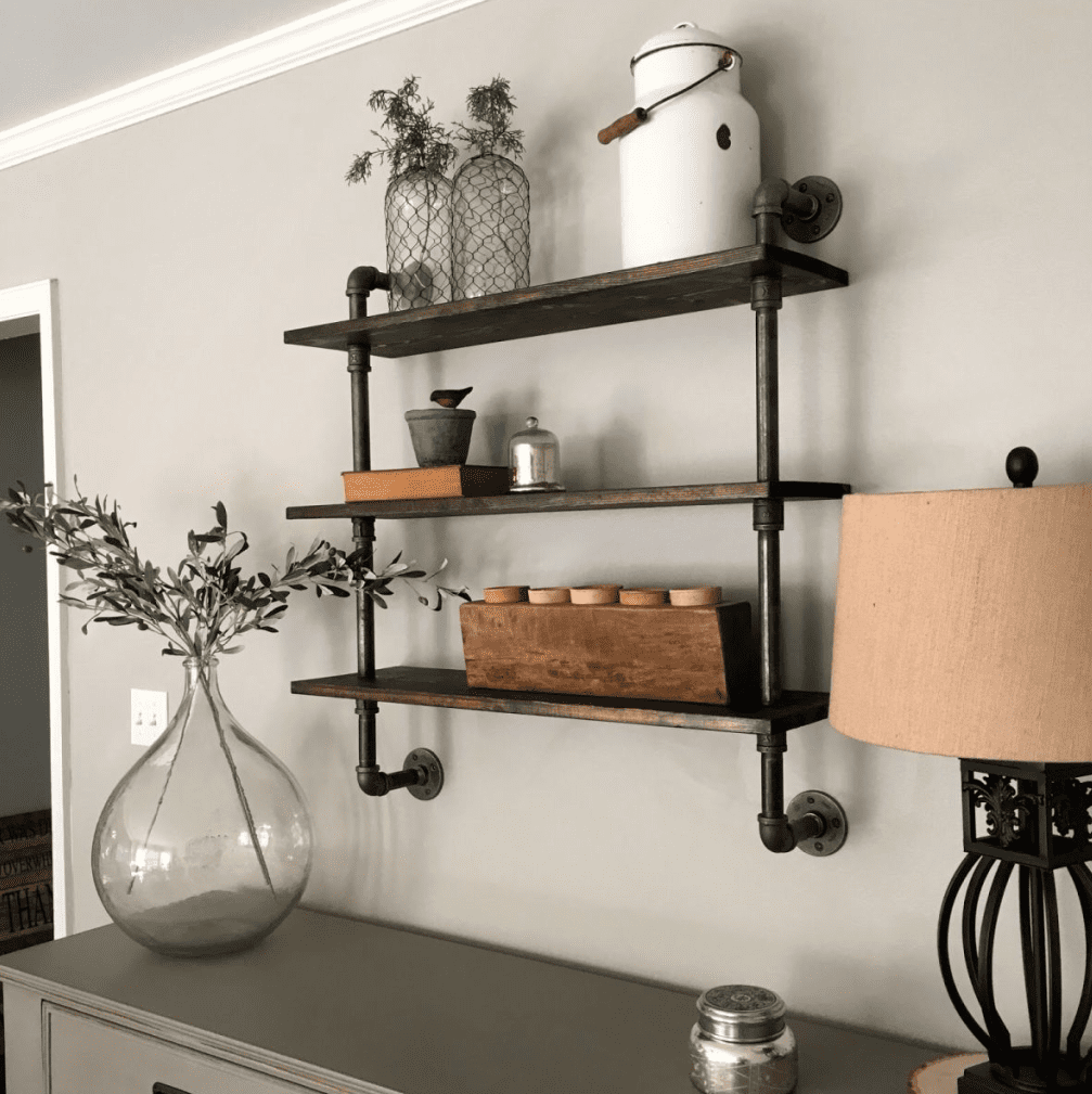 Wall mounted three tier shelf made from reclaimed metal pipes and dark wood boards, styled with plants, jars, and decor in a neutral living room