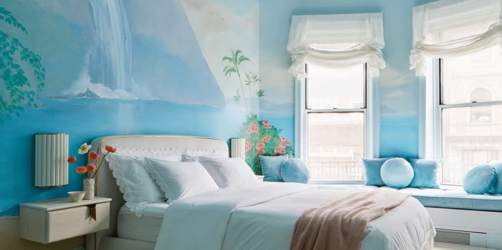 Light blue bedroom with a hand painted waterfall and tropical landscape mural behind the bed and window seat, white bedding, and pastel blue pillows.
