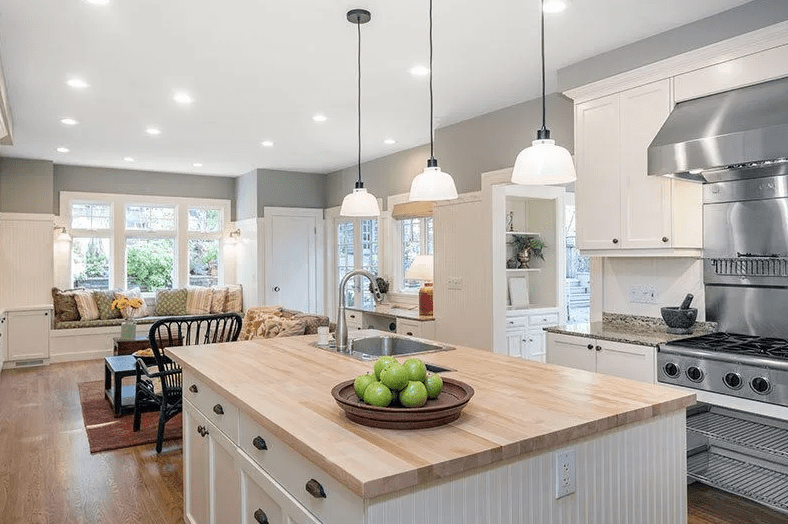 Bright open kitchen with a large wood island, three white pendant lights, stainless range, and a bowl of green apples as a simple centerpiece.