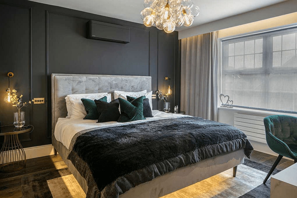 Modern master bedroom with dark walls, plush gray bed, emerald green accents, and layered lighting from pendant lamps and a glass bubble chandelier.