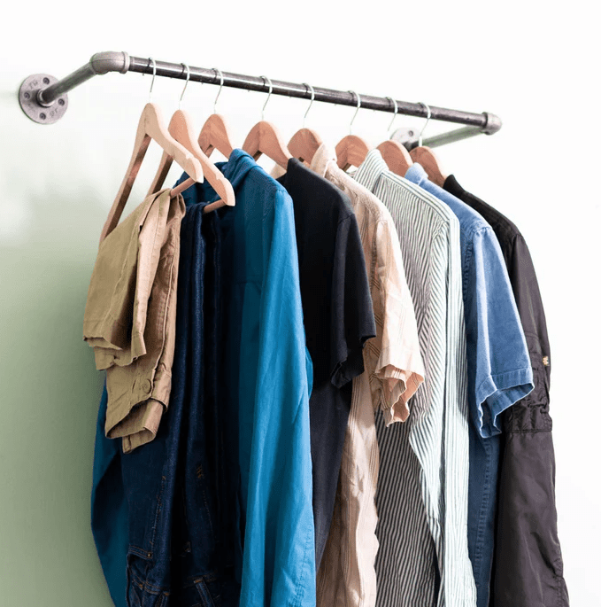 Wall mounted clothing rack made from reclaimed metal pipe with wooden hangers holding neatly arranged shirts and pants