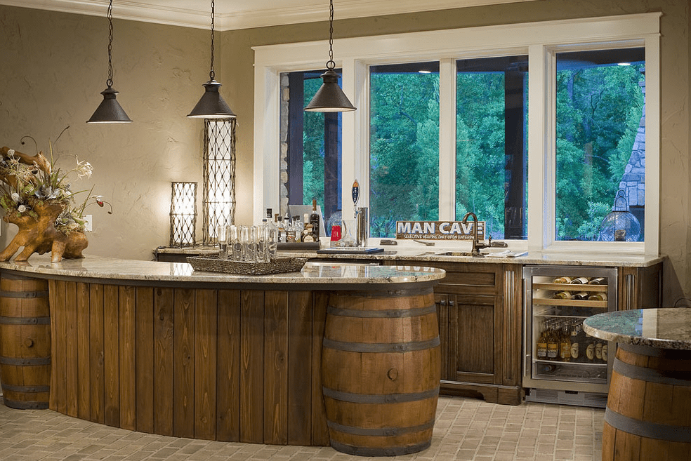 Home bar with a curved wood counter supported by large barrels, stone countertop, pendant lights, and a back wall of cabinets with a glass front mini fridge.