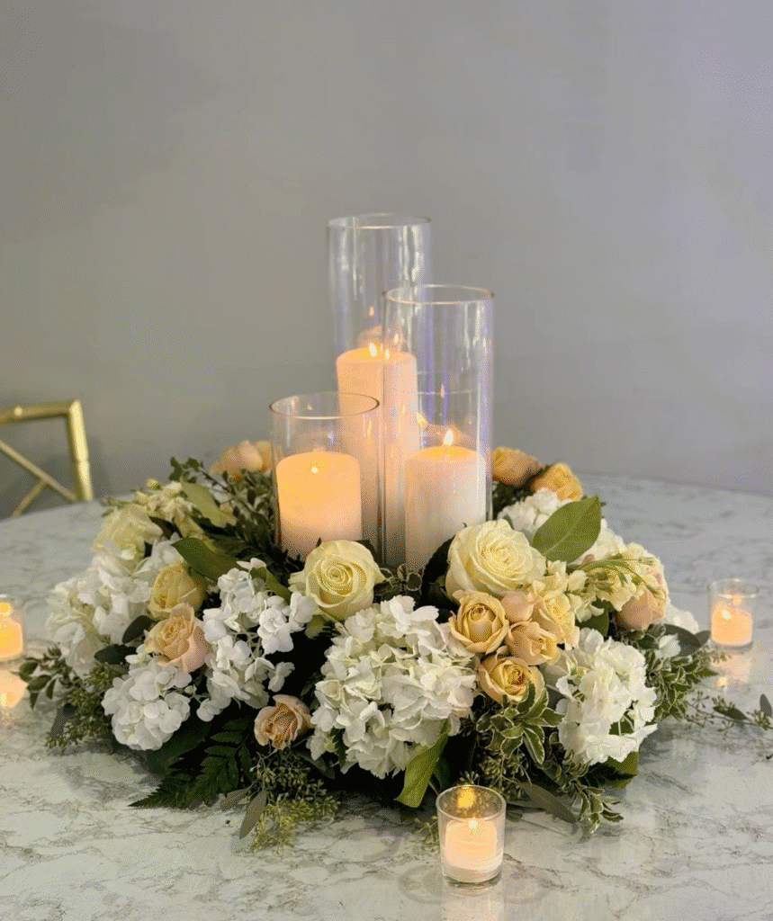 Low floral centerpiece of white hydrangeas and pale yellow roses with greenery, arranged around glowing pillar candles in clear glass cylinders on a marble tabletop.
