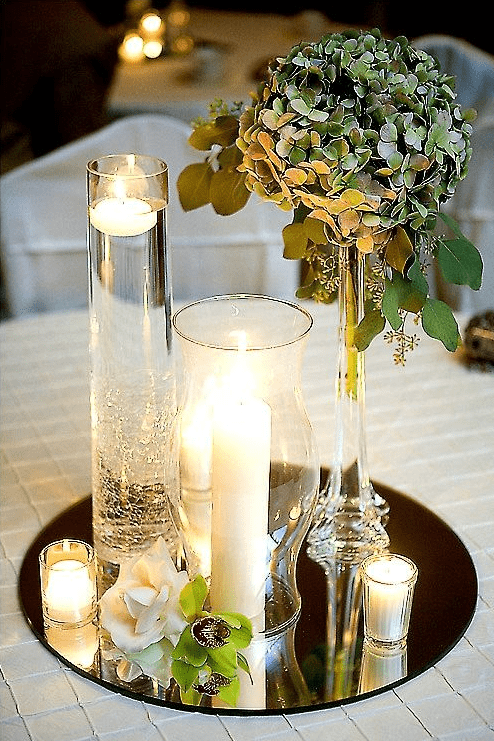 Round mirrored centerpiece with tall glass vases holding floating and pillar candles, a single hydrangea arrangement, and small votives all reflected in the mirror.