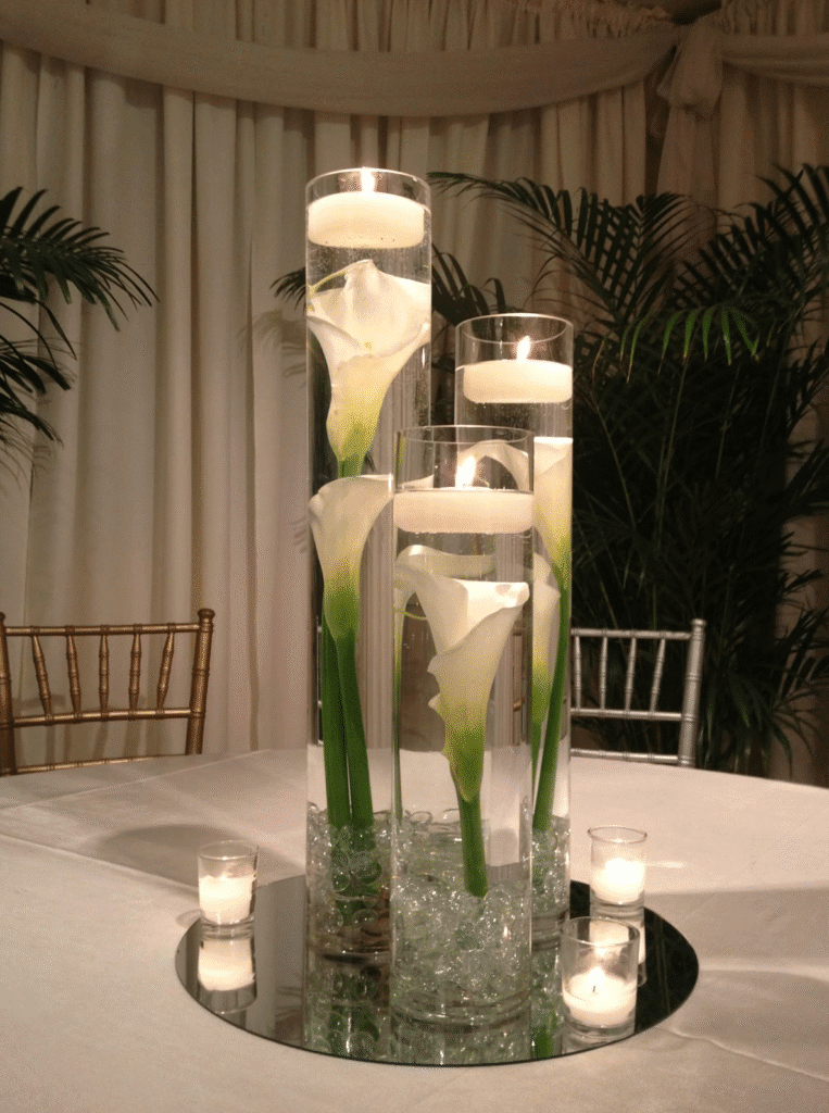 Tall glass cylinders filled with water and white calla lilies topped with floating candles arranged on a round mirror with small votives around the base.
