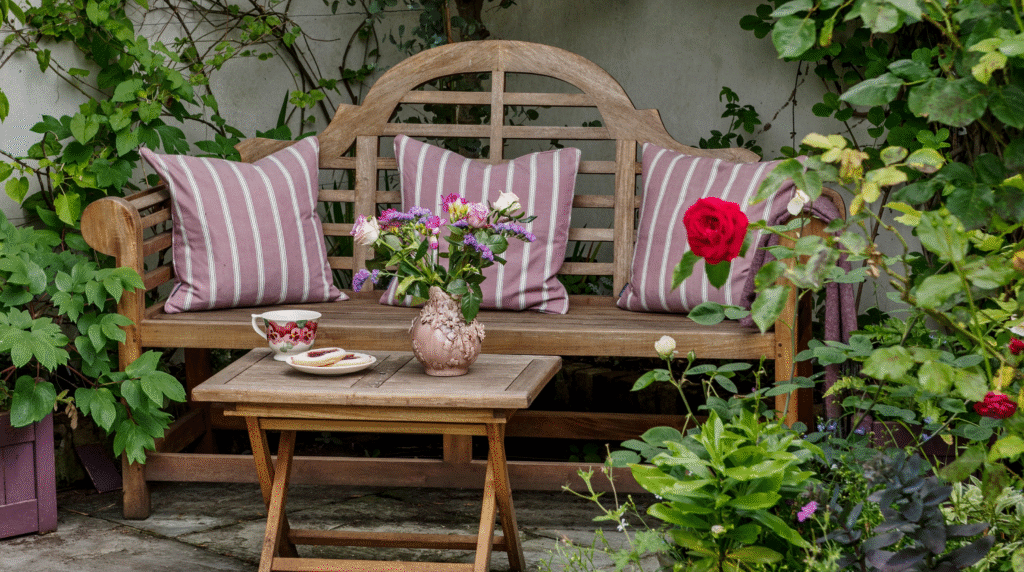 Cozy wooden garden bench with striped mauve cushions and a small rustic table holding a floral vase, teacup, and biscuits surrounded by lush greenery and roses.