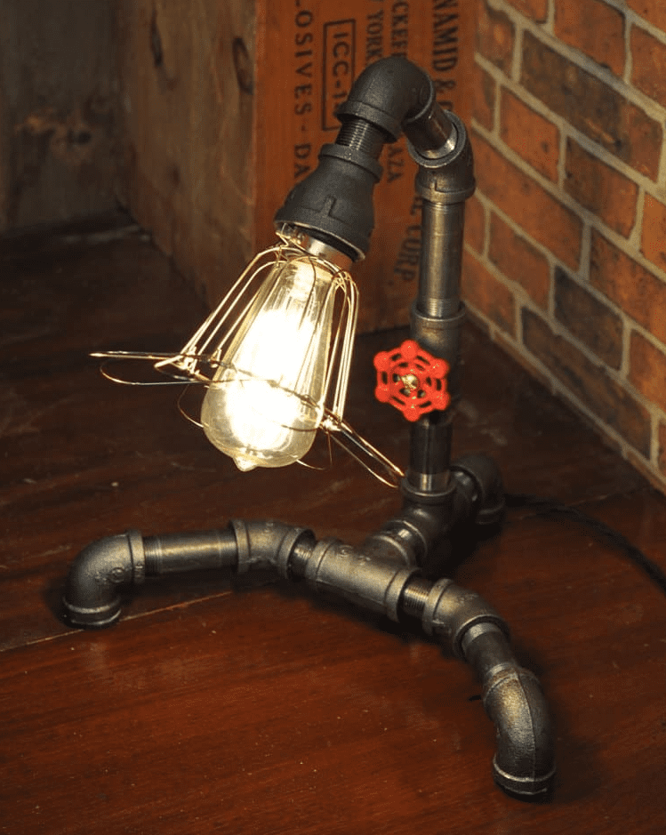 Industrial style table lamp made from dark reclaimed metal pipes with an exposed Edison bulb in a wire cage and a red valve handle detail on a wooden surface