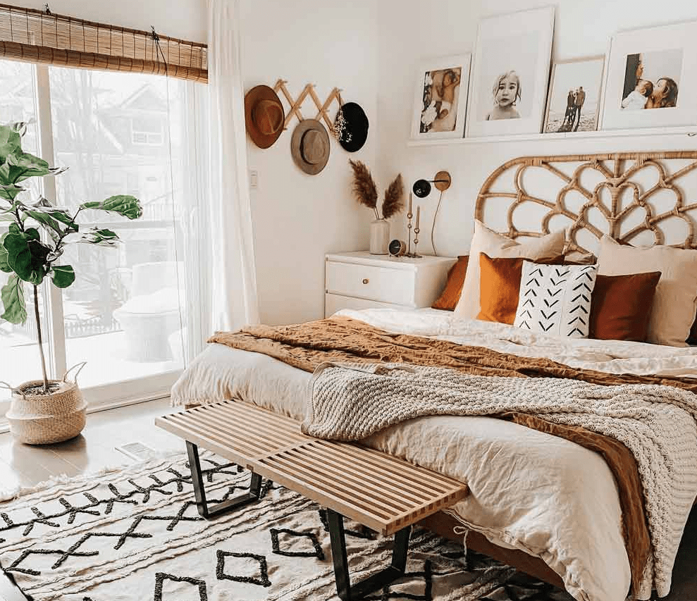 Cozy bohemian bedroom with rattan headboard, layered neutral bedding, warm rust pillows, wall art above the bed, and a wooden bench on a patterned rug.
