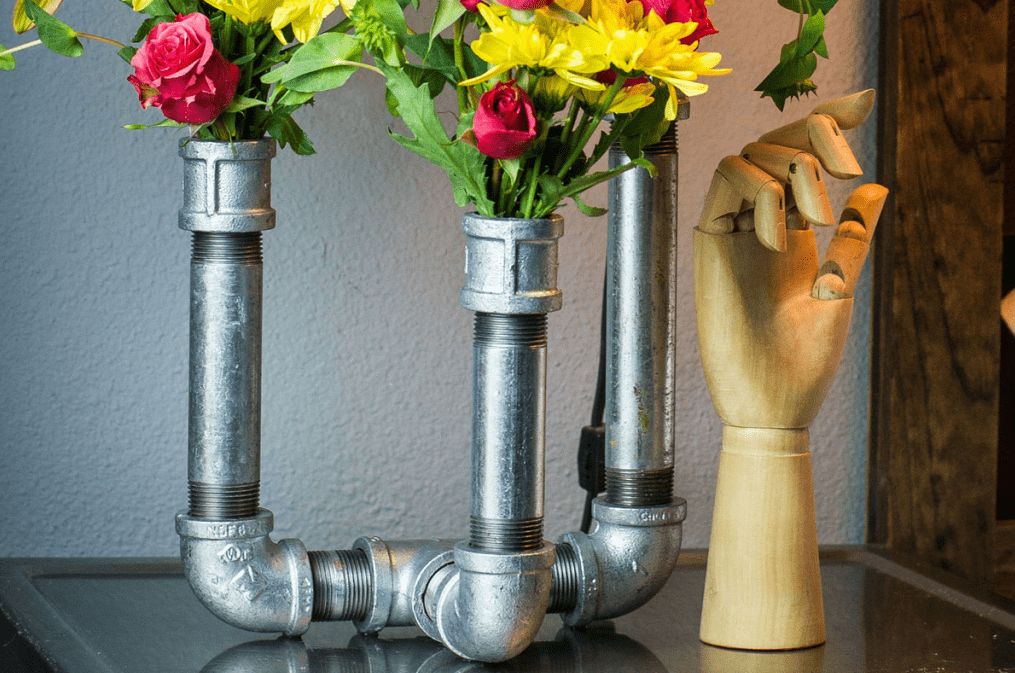 Upcycled metal plumbing pipes turned into a vase holding bright pink and yellow flowers with a wooden mannequin hand beside it on a tabletop