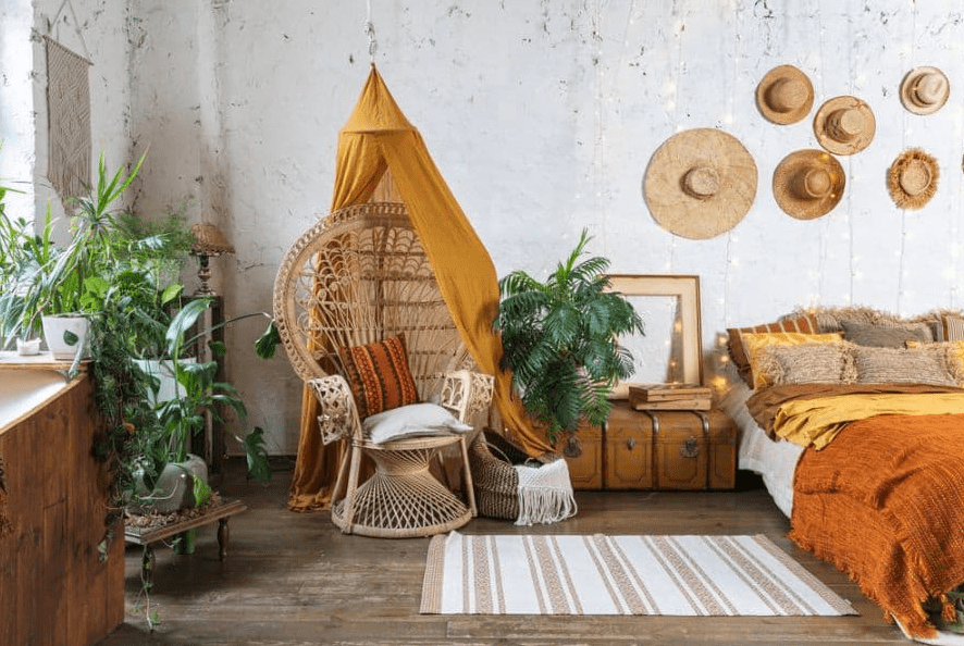 Bohemian bedroom corner with rattan peacock chair under mustard canopy, lush green plants, vintage trunks, and straw hats on a textured white wall.