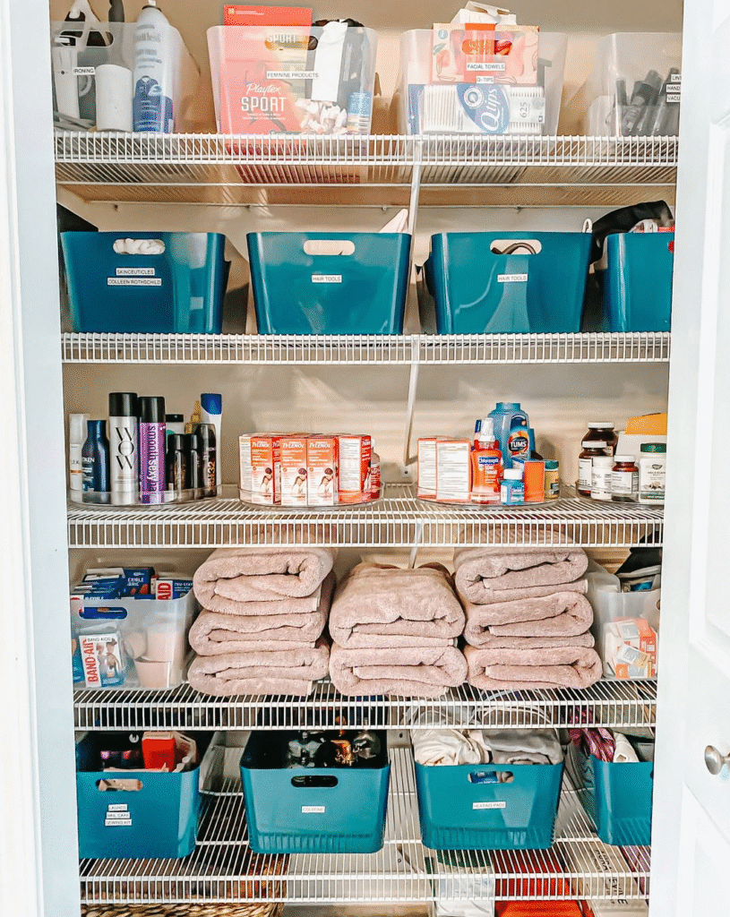 Organized linen closet with teal storage bins, labeled containers, folded beige towels, and neatly arranged toiletries and medicines on white wire shelves.