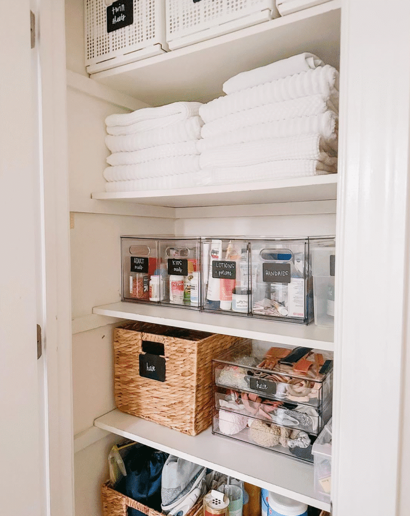 Organized linen closet with stacks of white towels, clear labeled bins for toiletries, woven baskets, and pull out organizers neatly arranged on white shelves.