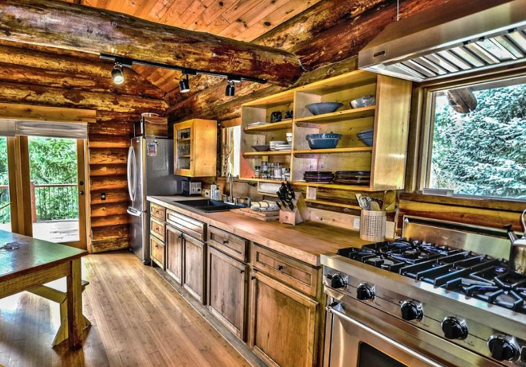 Cozy rustic log cabin kitchen with reclaimed barn wood cabinets, long butcher block counters, open shelves, and stainless steel appliances facing a forest view.