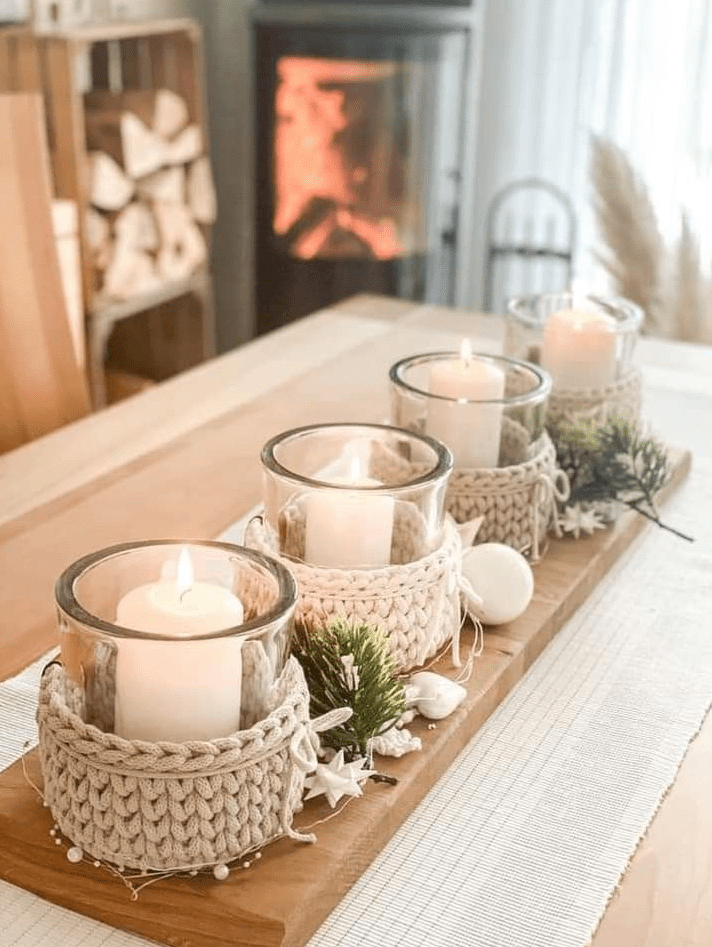 Row of glass candles in cream crochet upcycled sleeves on a wooden tray, decorated with greenery on a cozy table near a fireplace.