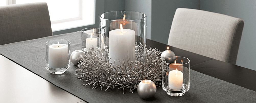 Modern dining table with white pillar candles in clear glass holders, silver branch style wreath, and metallic ornaments arranged along a gray runner.