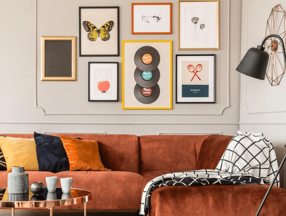 Cozy bedroom or lounge with a rust colored sectional sofa, colorful pillows, and a gallery wall of framed art and prints including a butterfly, apple, records, and abstract line drawings.