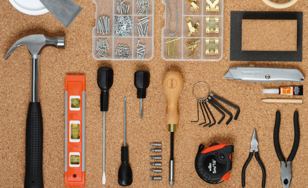 Assorted homeowner tools laid out on a corkboard, including a hammer, level, screwdrivers, tape measure, pliers, utility knife, hex keys, and hardware