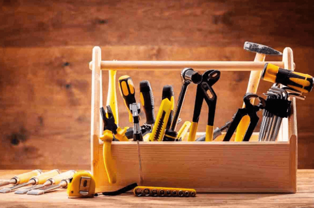 Wooden toolbox filled with basic hand tools like screwdrivers, pliers, a utility knife, and a tape measure on a workbench