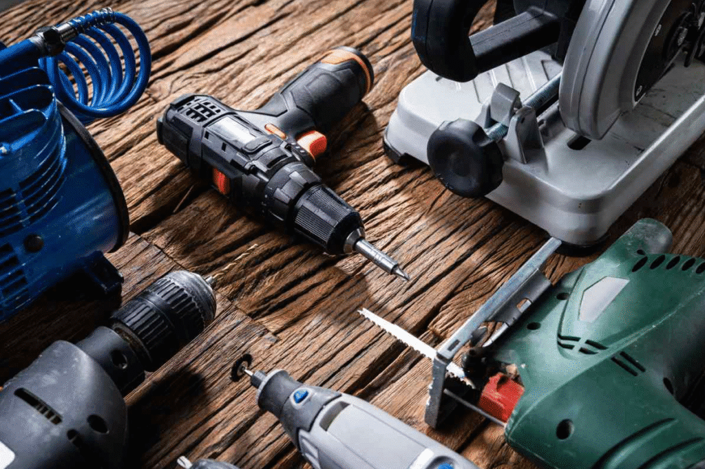 Assorted power tools on a wooden surface, including a cordless drill, jigsaw, rotary tool, and other workshop equipment