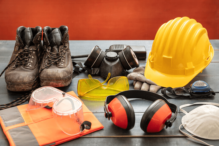 Home safety gear on a table including a hard hat, safety goggles, ear protection, dust mask, gloves, work boots, and a respirator