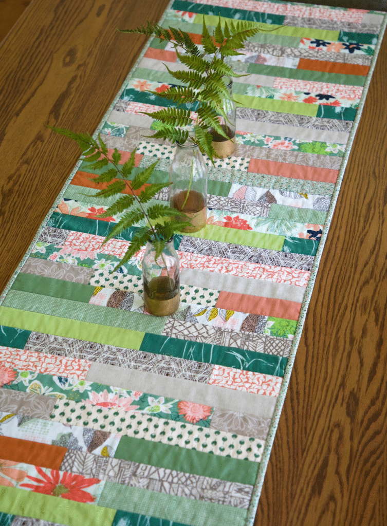 Patchwork table runner made from fabric scraps in green, coral, and neutral prints, styled with small glass vases of fern stems