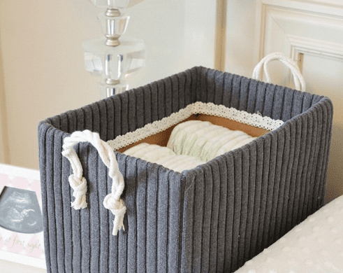 Rectangular storage basket covered in a ribbed gray sweater fabric with rope handles holding rolled baby diapers on a white nursery dresser