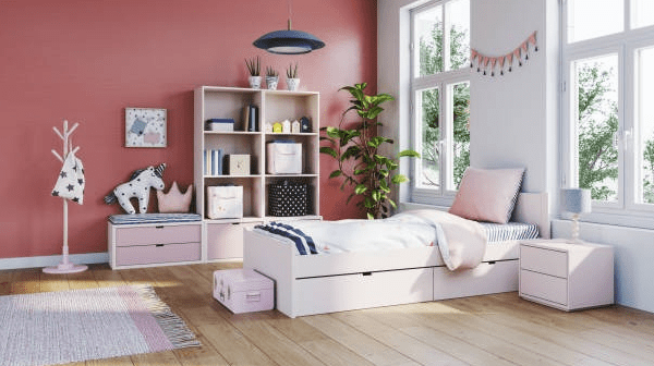 Bright child bedroom with a soft coral statement wall, white bed with storage drawers, pink rug, shelves with toys, and large windows letting in natural light