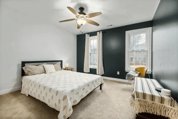 Bedroom with dark green accent walls, white trim, beige carpet, simple bed with patterned duvet, wooden side table, striped bench, and ceiling fan