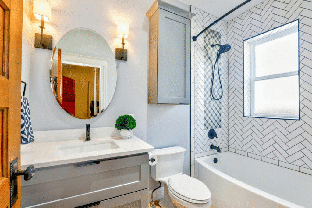 Small modern bathroom with gray vanity, round mirror, warm wall sconces, and white tub surrounded by patterned tile and dark fixtures showing stylish updates on a budget