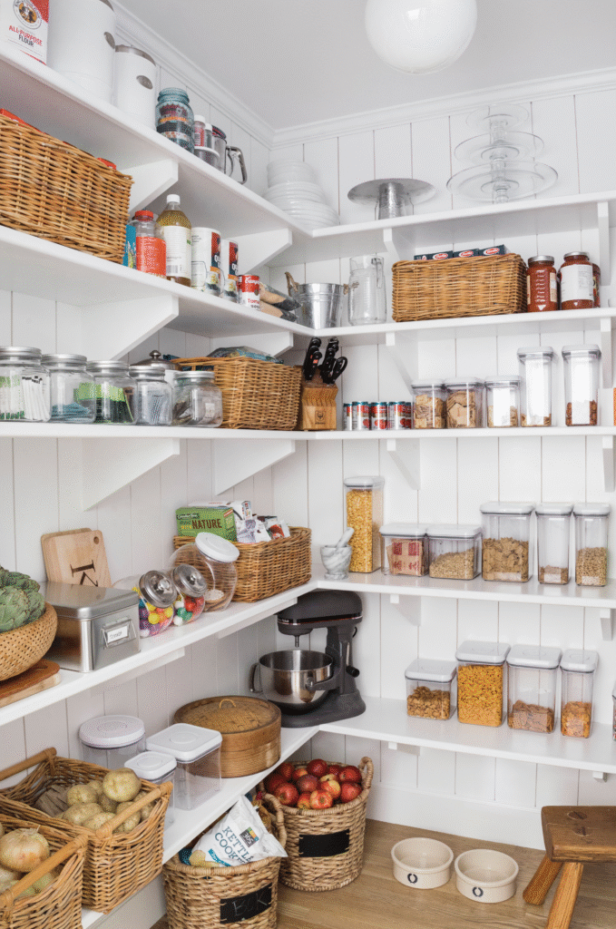 Organized white pantry with open shelves, clear food containers, woven baskets, and crates holding produce and snacks