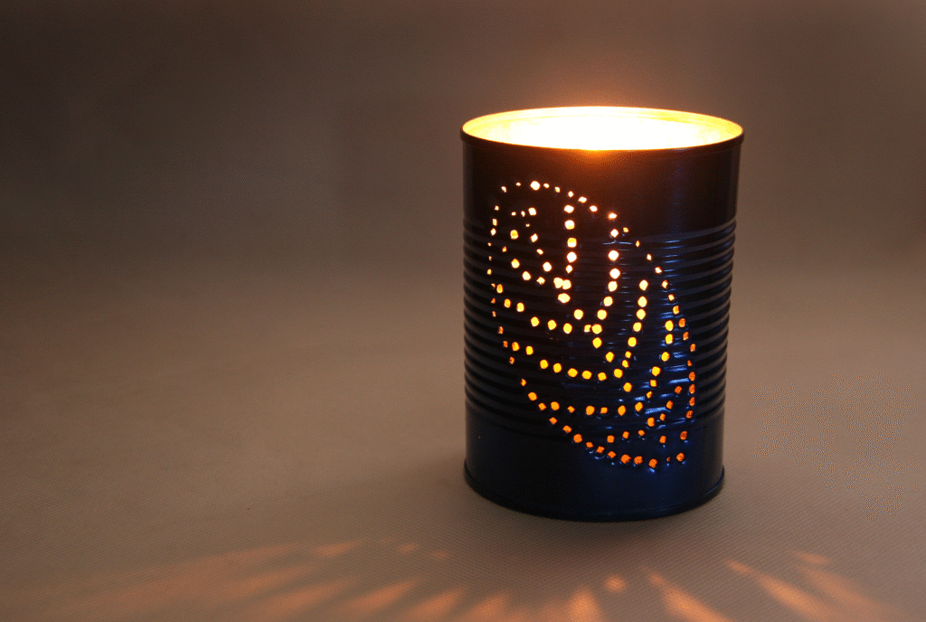Glowing upcycled tin can lantern with a spiral pattern of punched holes casting warm light on the surface in a dark room